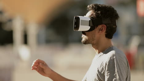Side-view-of-young-Arabic-man-with-dark-curly-hair-and-beard-in-grey-T-shirt-being-in-park-in-virtual-reality-glasses